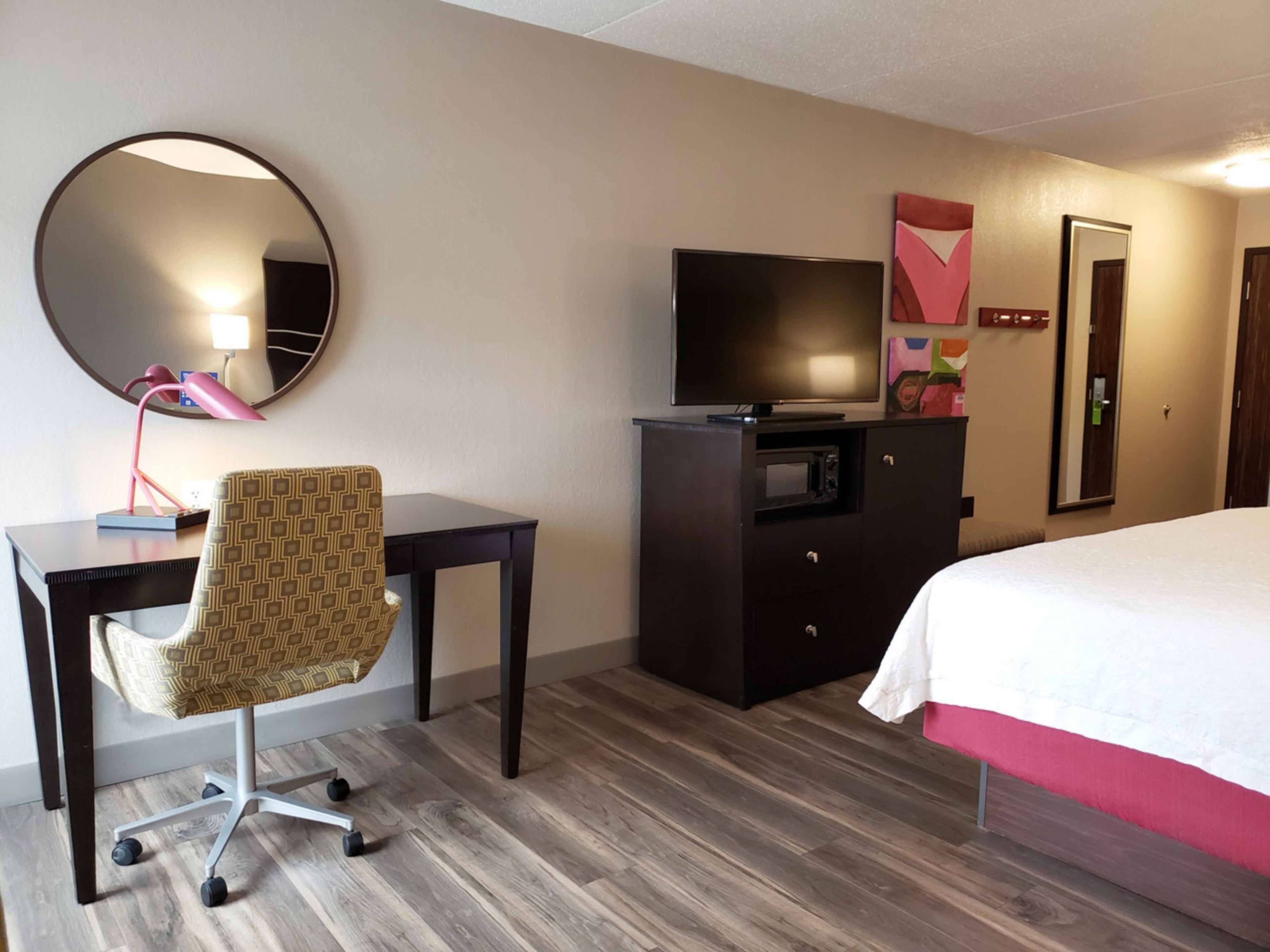 A hotel room with a bed, a desk with a chair, a TV, a microwave, and a mirror.
