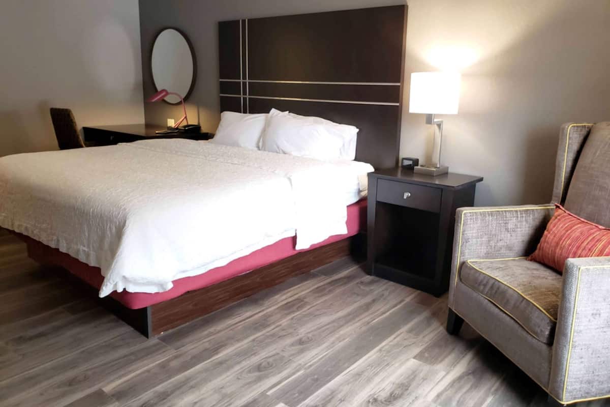 A hotel room with a large bed, a nightstand with a lamp, a chair, and a wall-mounted mirror.