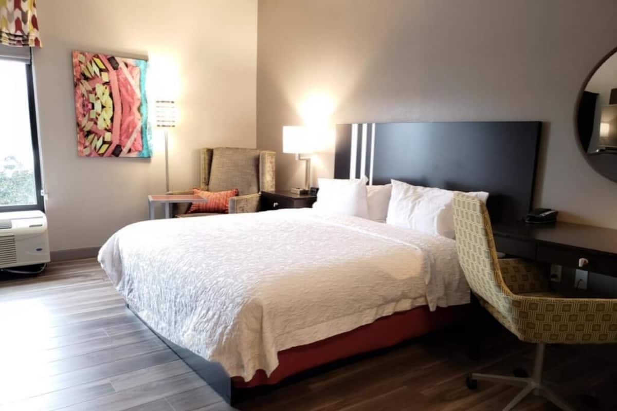 A hotel room with a large bed, a chair, a desk, a lamp, and a painting on the wall.