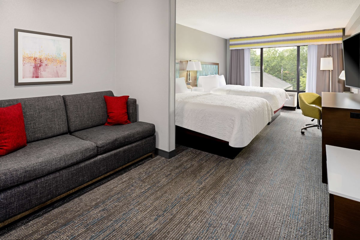 A hotel room with a gray sofa, two beds, a desk, and a TV.
