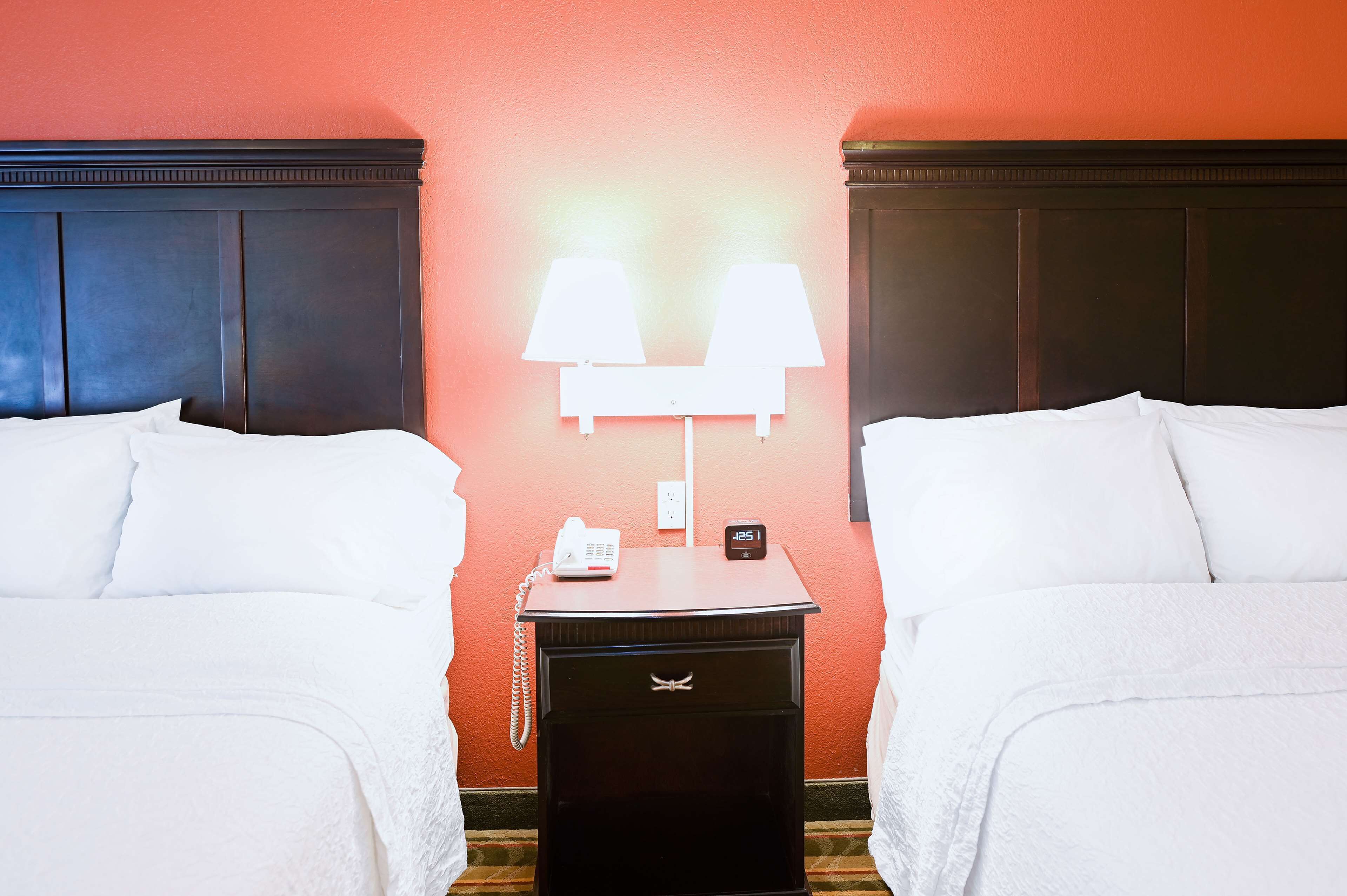 A hotel room with two beds, a nightstand with a lamp, a phone, and a clock.