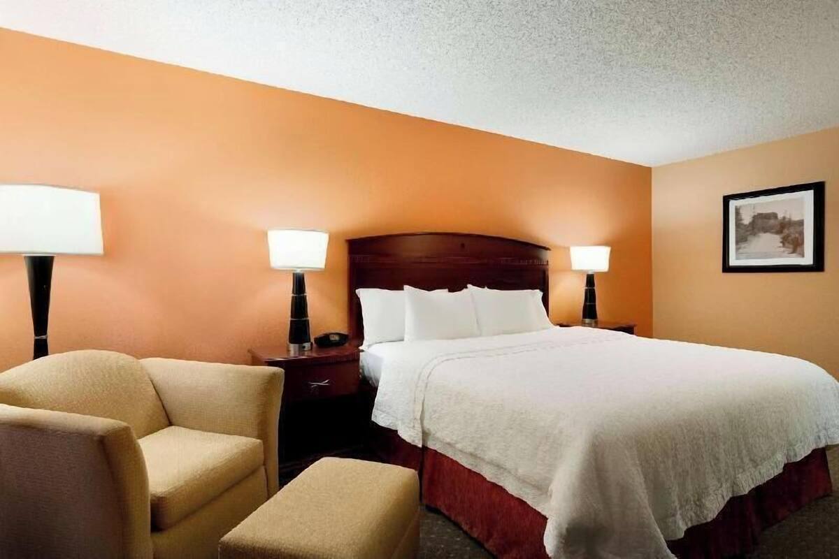A hotel room with a bed, two bedside lamps, a chair, a nightstand, and a framed picture on the wall.