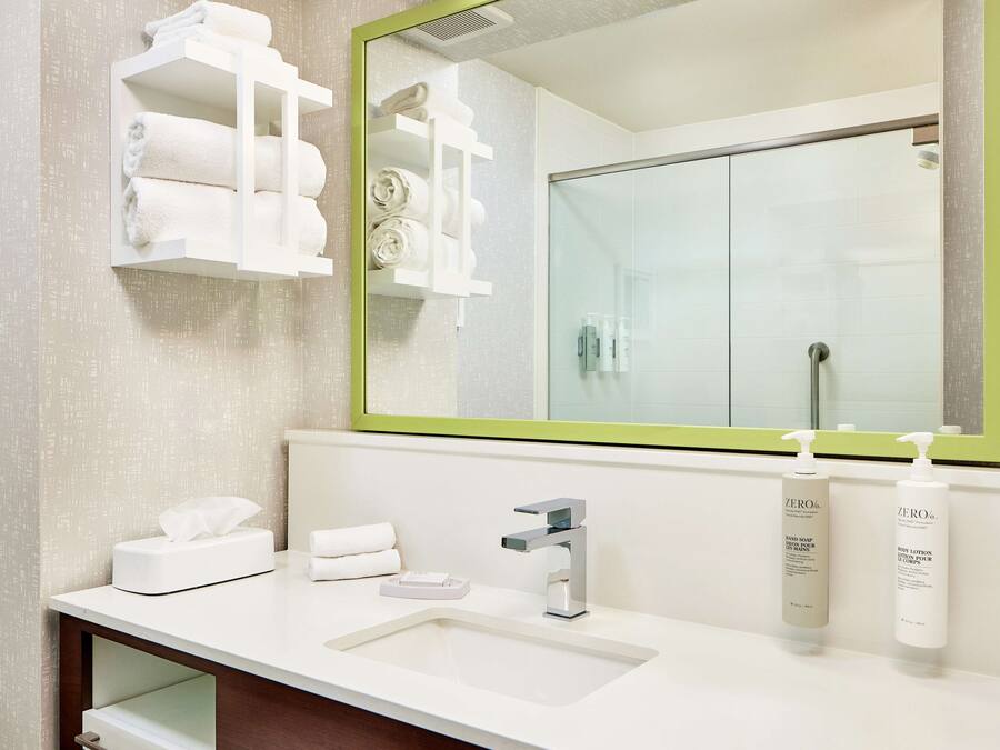 A modern bathroom with a white sink, a large mirror with a green frame, and shelves holding towels and toiletries.