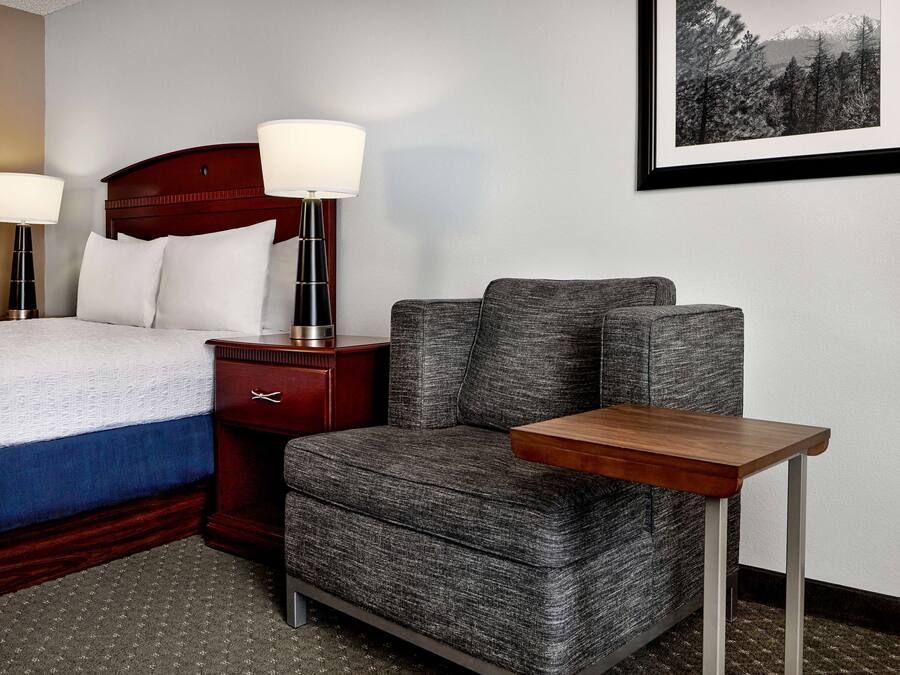 Habitación de hotel con cama, una silla tapizada en gris, una mesita de noche de madera y un cuadro en la pared.