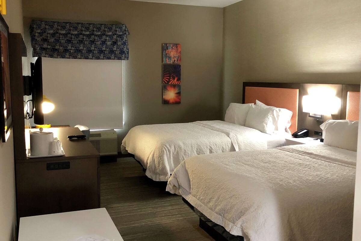 A hotel room with two beds, a desk with a lamp, a telephone, and a wall-mounted TV.