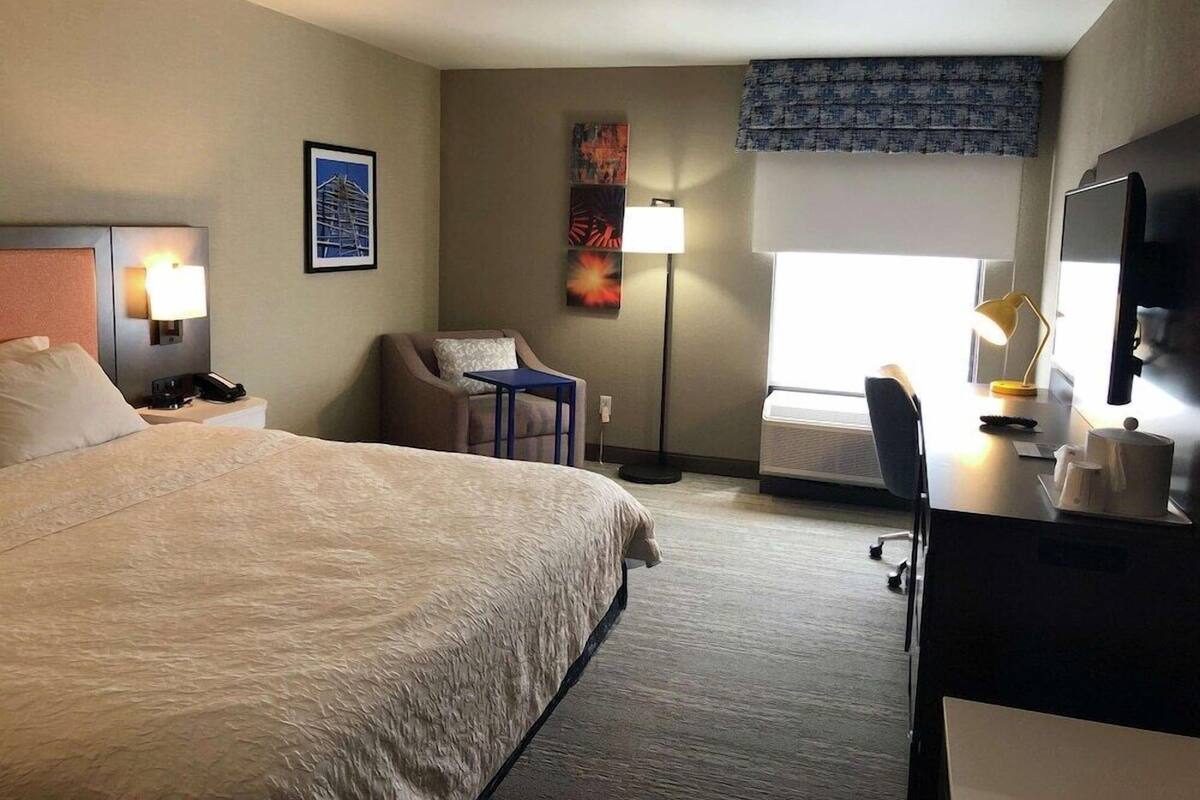 A hotel room with a bed, desk, chair, lamp, artwork, and a window with blinds.