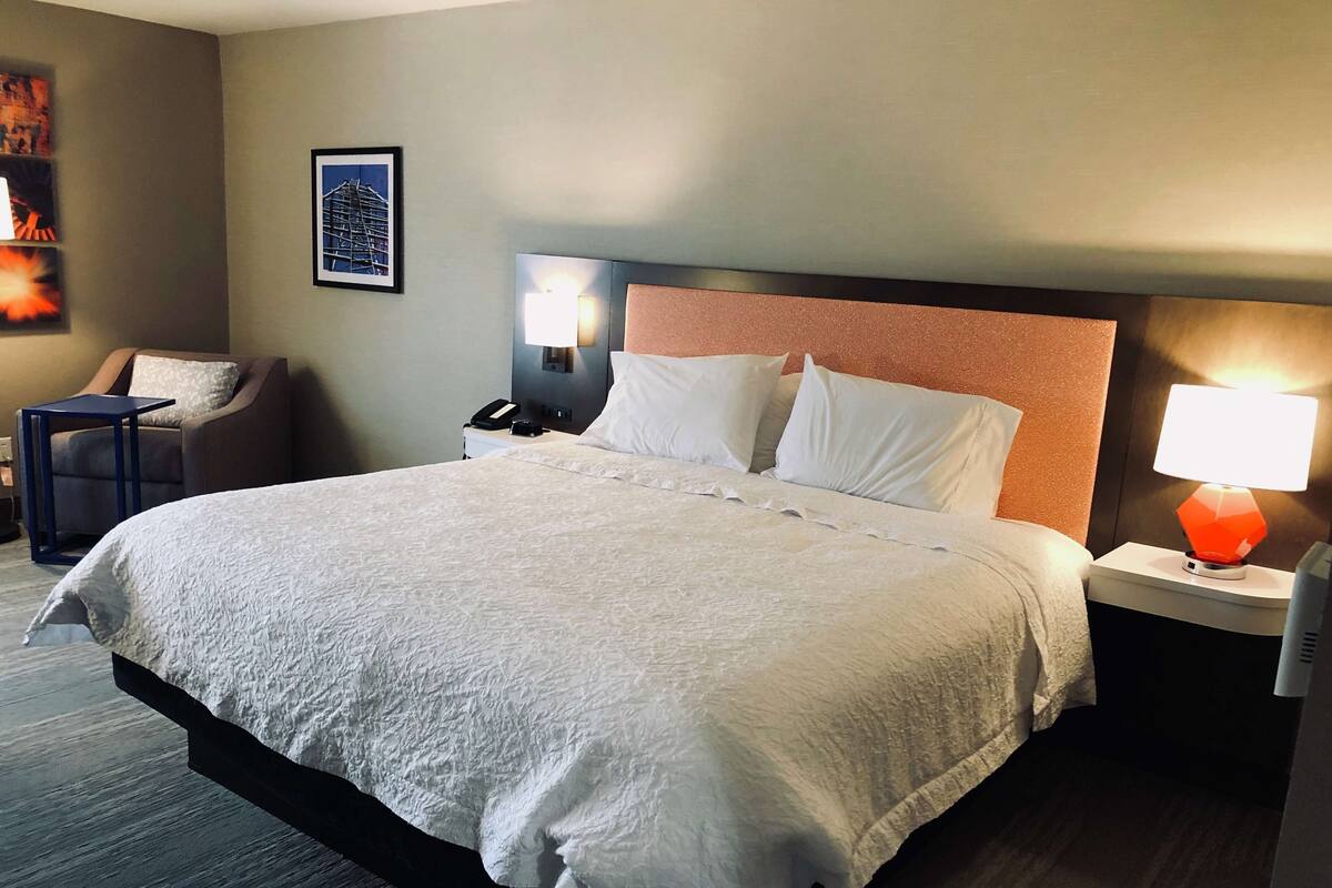 A hotel room with a large bed, a nightstand with a lamp, a chair, and a framed picture on the wall.