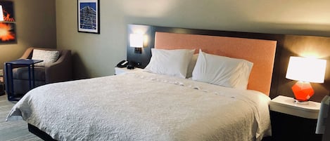 A hotel room with a large bed, a nightstand with a lamp, a chair, and a framed picture on the wall.