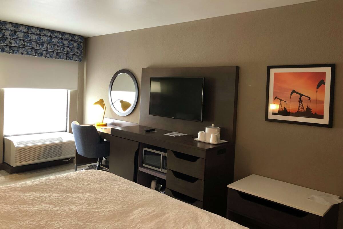 A hotel room with a bed, desk, chair, TV, and a painting of an oil rig.