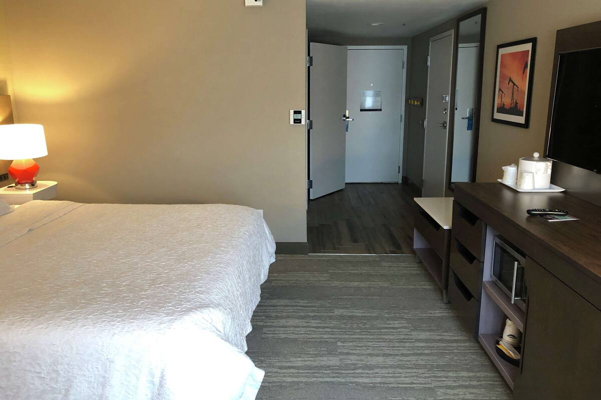 A hotel room with a bed, a desk with a lamp, a television, and a bathroom visible through an open door.