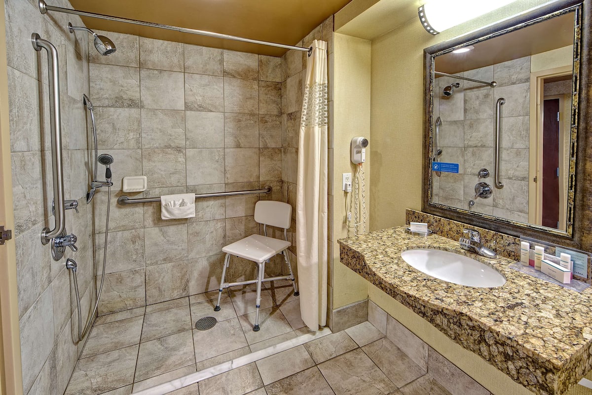 A bathroom with a shower, grab bars, a white chair, a sink, and a mirror.