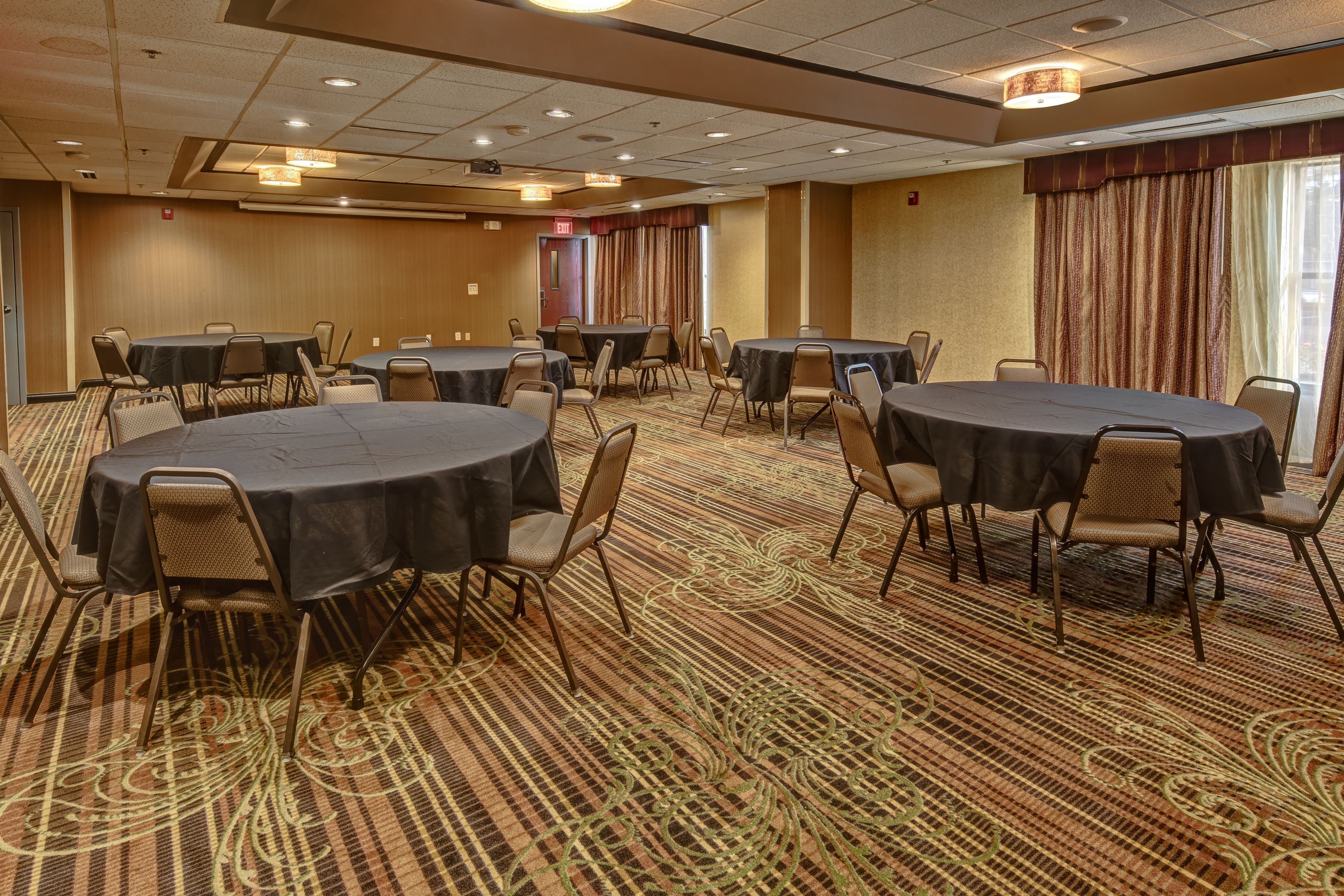 A conference room with multiple round tables and chairs, carpeted flooring, and a large window with curtains.