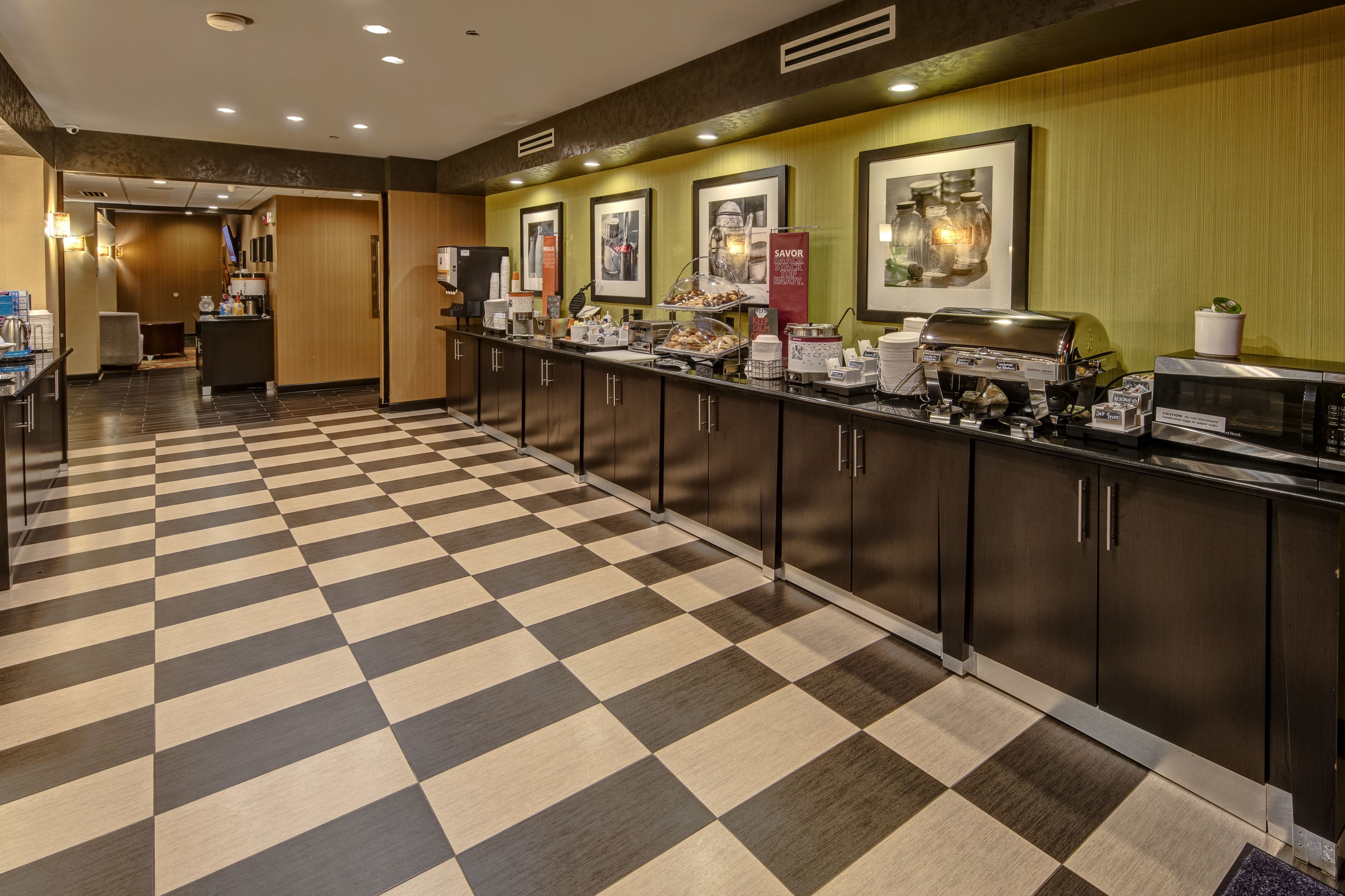 Una cocina de hotel con piso a cuadros, un mostrador de buffet con diferentes alimentos y fotografías enmarcadas en la pared.