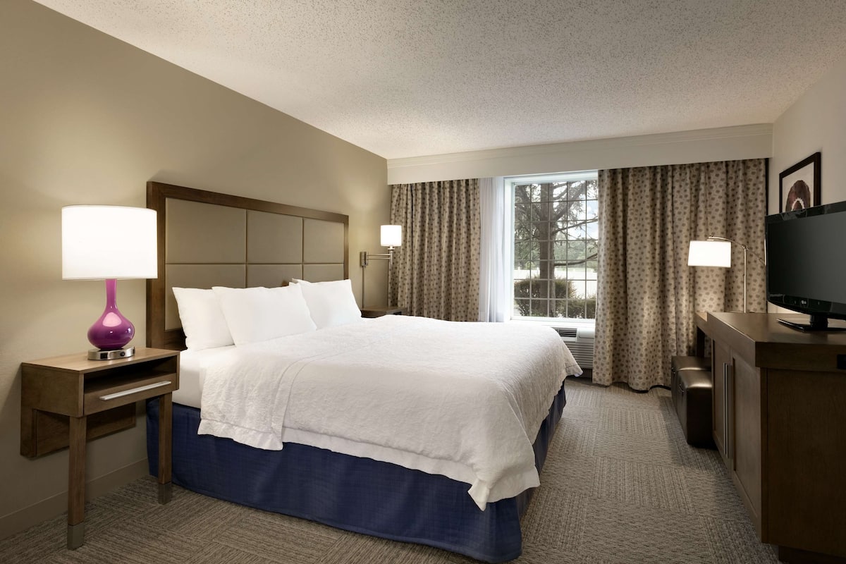 A hotel room with a large bed, a desk with a television, a lamp, and a window with curtains.