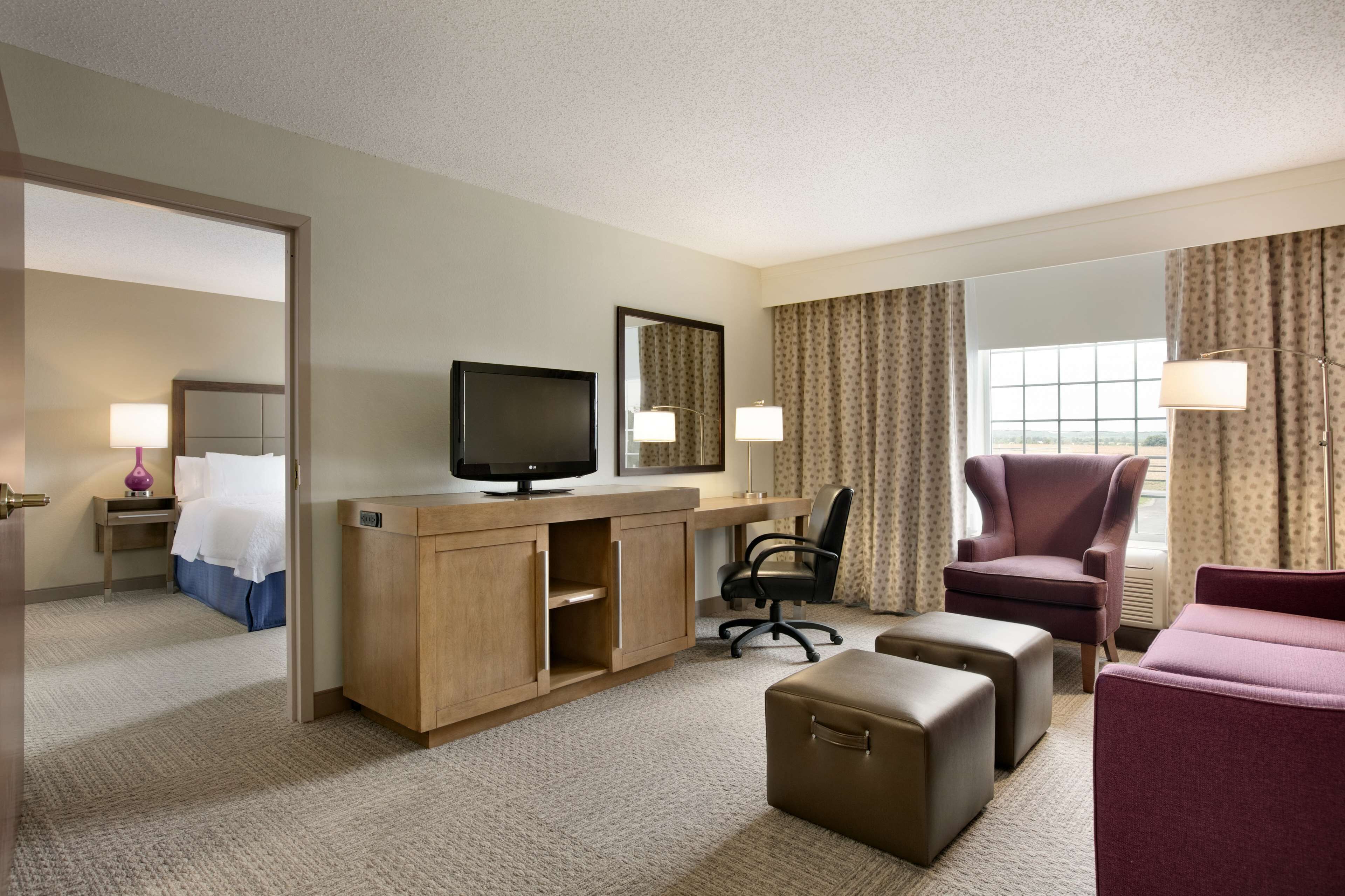 A hotel room with a bed, desk, TV, chair, ottoman, and curtains.
