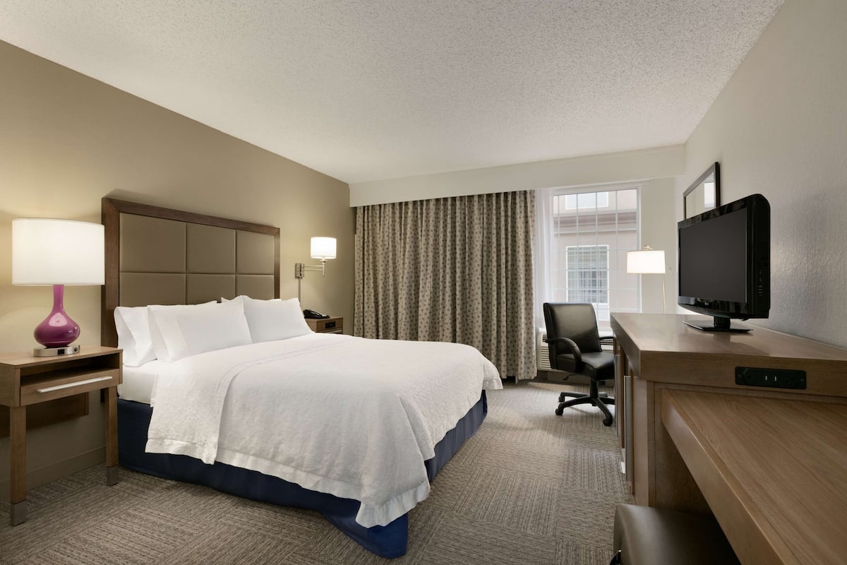 A hotel room with a bed, a desk with a television, a chair, a lamp, and a window with curtains.