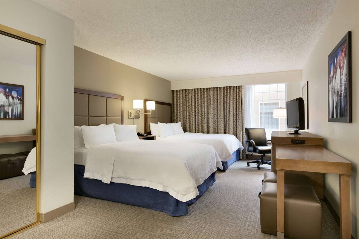 A hotel room with two beds, a desk, a chair, a TV, and a window with curtains.