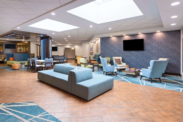 A spacious lobby with a grey sectional sofa, multiple armchairs, a coffee table, and a flat-screen TV mounted on the wall.