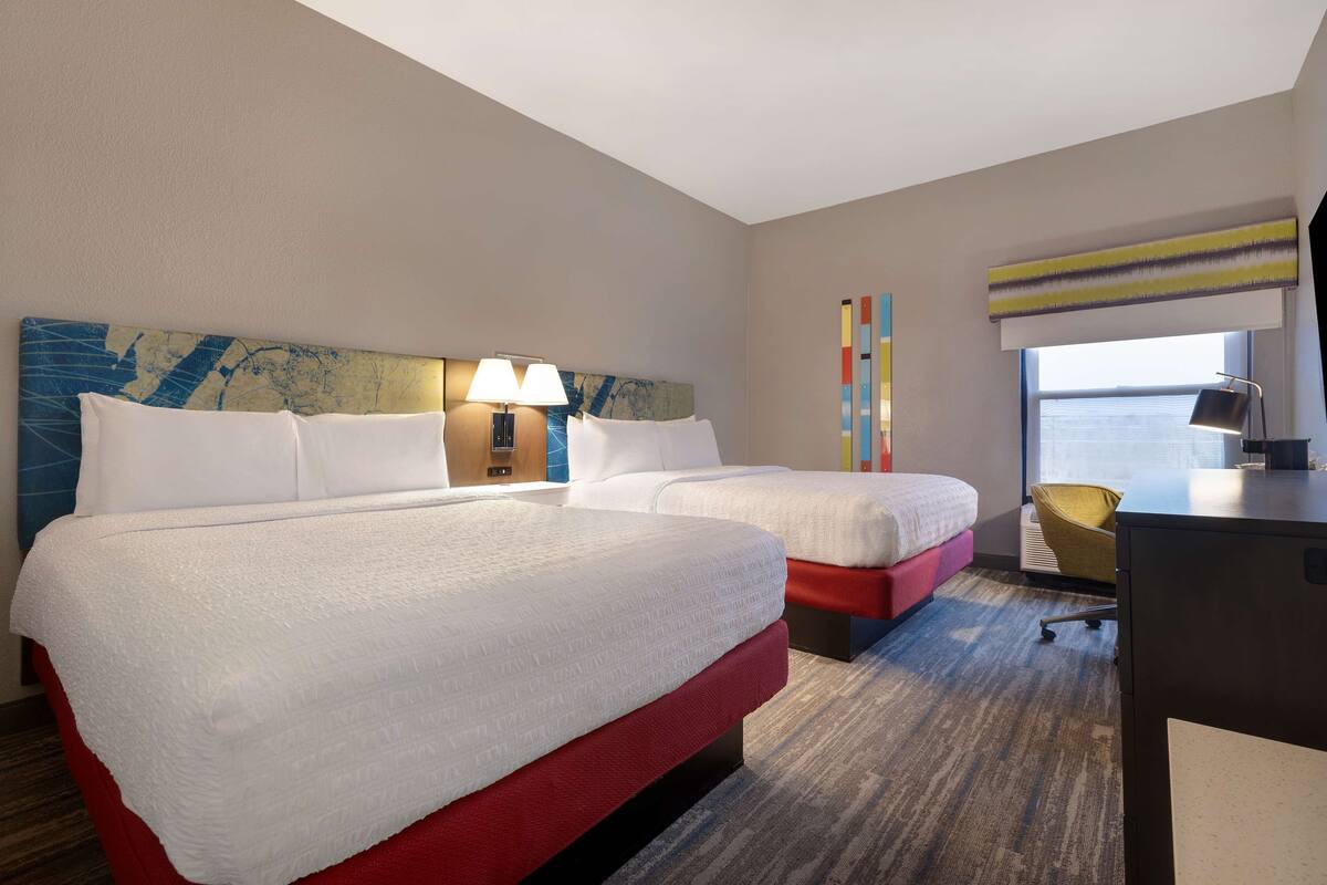 A hotel room with two beds, a desk, a chair, and a window with a view.
