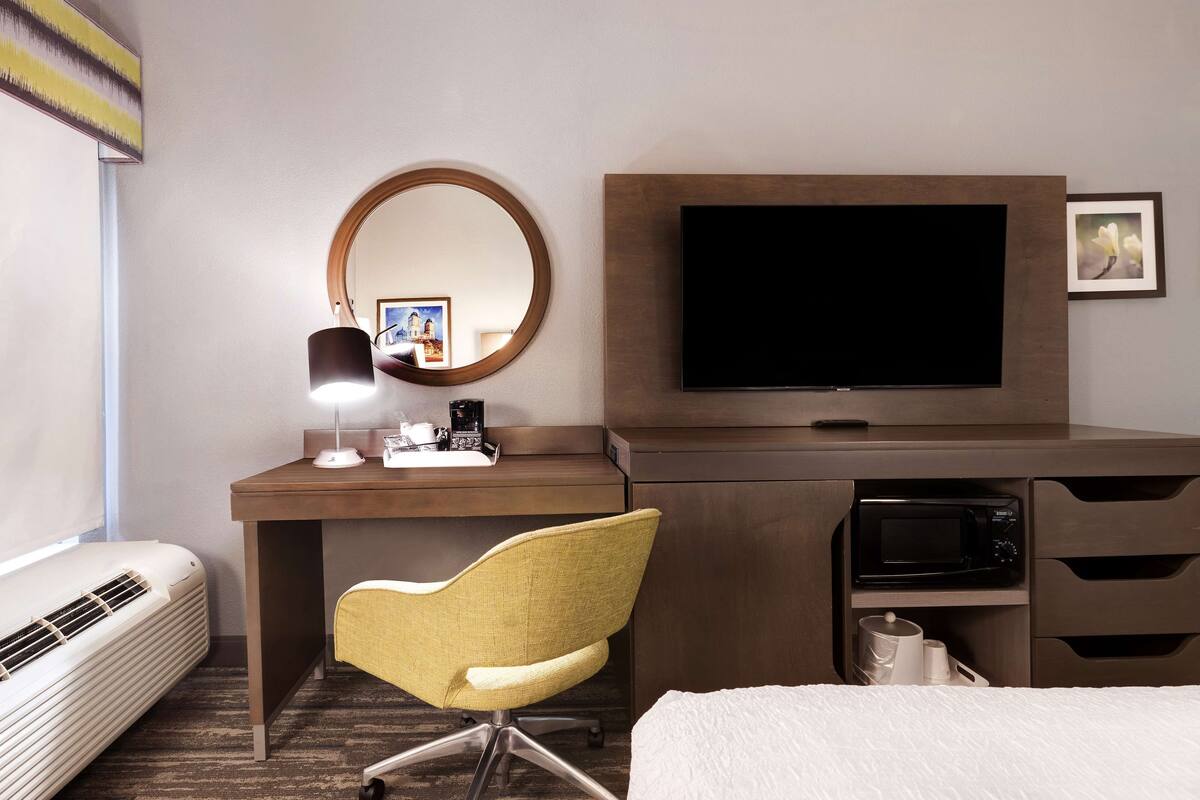 A hotel room with a bed, a desk with a computer, a chair, a TV, a microwave, and a mirror.