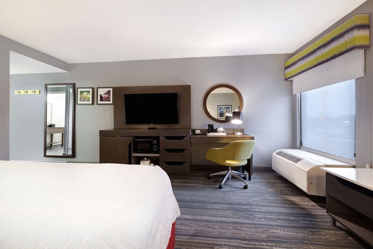A hotel room with a bed, a desk with a chair, a television, a mirror, and a window.