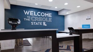 Un espace d’accueil avec un mur bleu sur lequel est affiché le message « BIENVENUE DANS L’ÉTAT CRÉOLE » et un panneau indiquant la présence d’un « responsable de service ». On y trouve deux bornes de libre-service et un panneau « Hilton Honors ».