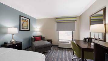 A hotel room with a bed, a desk, a chair, a nightstand, a lamp, a window with a view, and a painting on the wall.