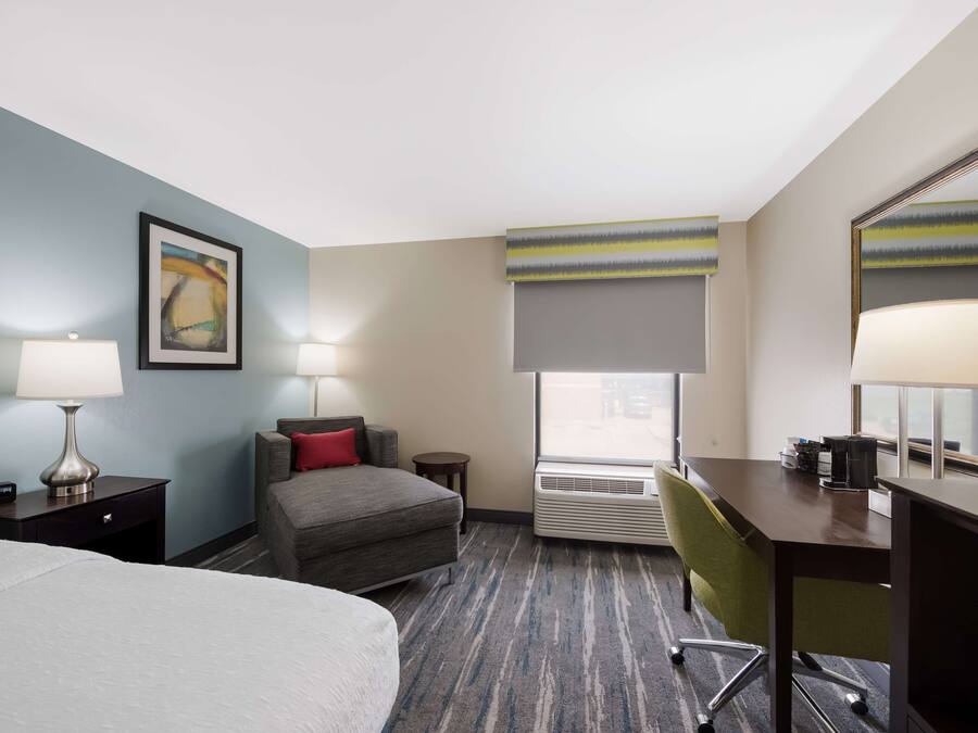 A hotel room with a bed, a desk, a chair, a nightstand, a lamp, a window with a view, and a painting on the wall.