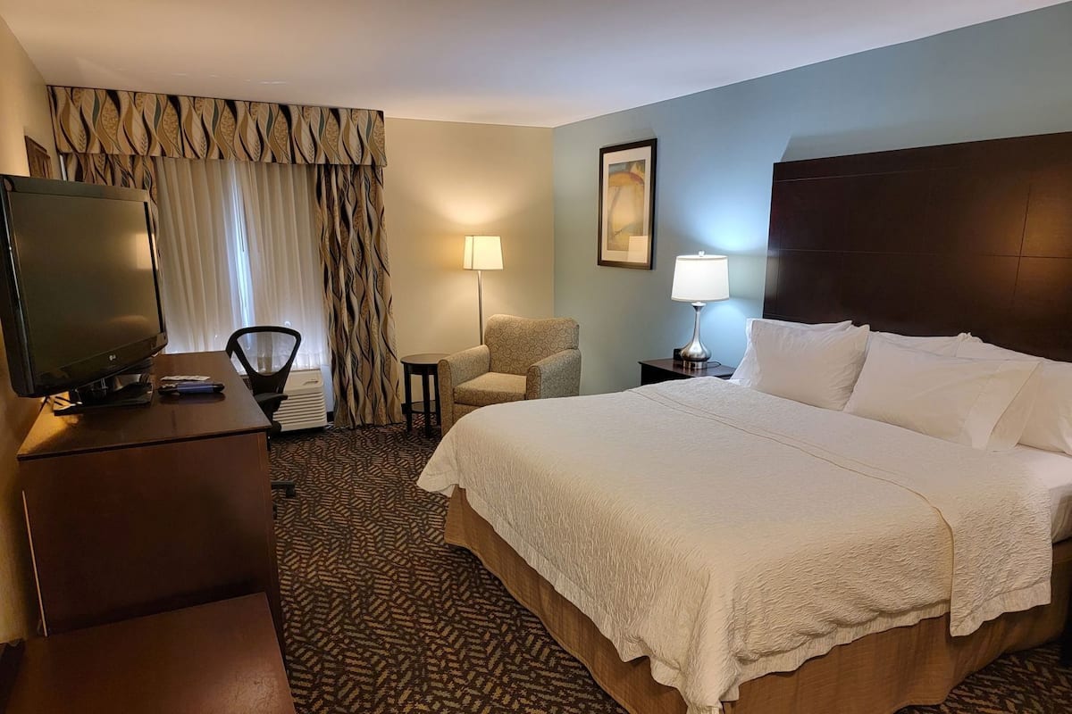 Habitación de hotel con una cama grande, un escritorio con televisor, una silla, una lámpara y un cuadro en la pared.