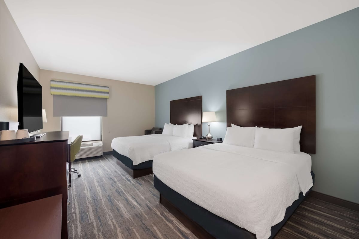 Habitación de hotel con dos camas, un escritorio, una silla y un ventanal grande.