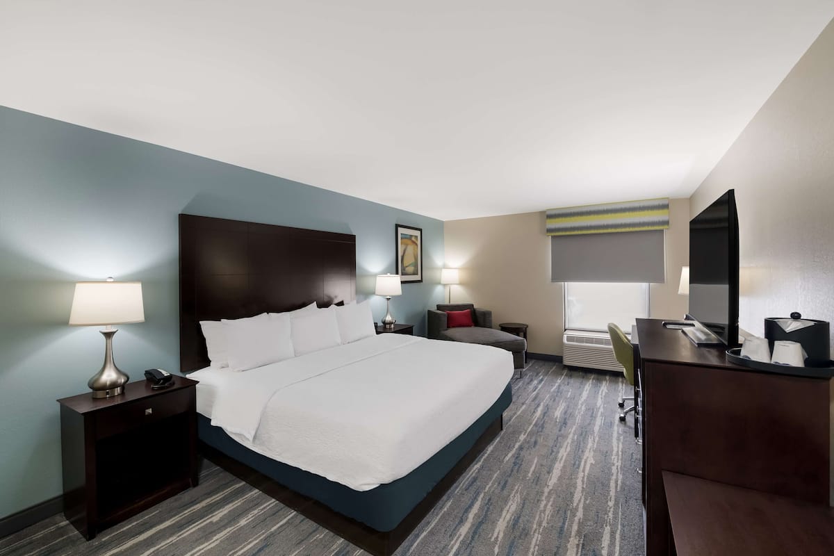 Habitación de hotel con una cama grande, mesitas de noche con lámparas, un escritorio con silla y un ventanal con persianas.