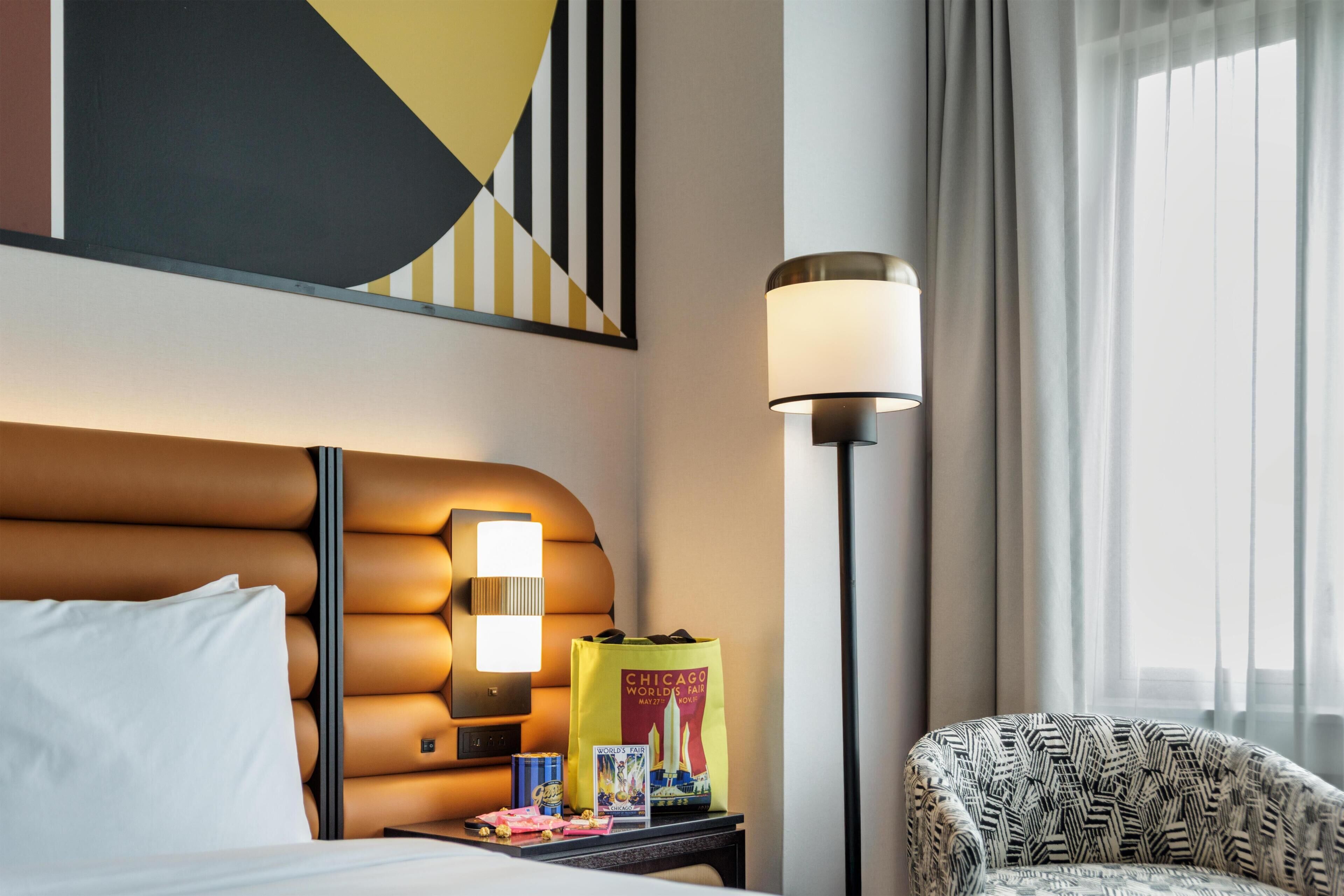 A hotel room with a bed, a nightstand with a lamp, a chair, a wall art piece, and a window with curtains.