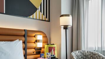 A hotel room with a bed, a nightstand with a lamp, a chair, a wall art piece, and a window with curtains.
