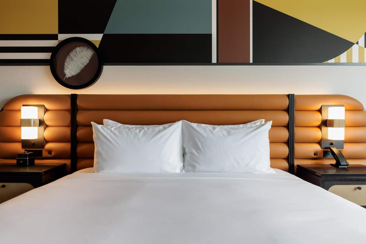 A hotel room with a bed, two bedside lamps, a wall clock, and a geometric wall design.