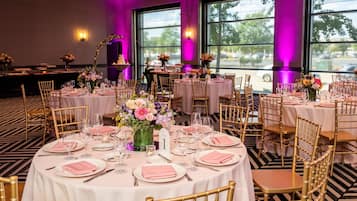 Een feestzaal met ronde tafels, ingericht voor een formeel evenement, met gouden stoelen en witte tafelkleden.