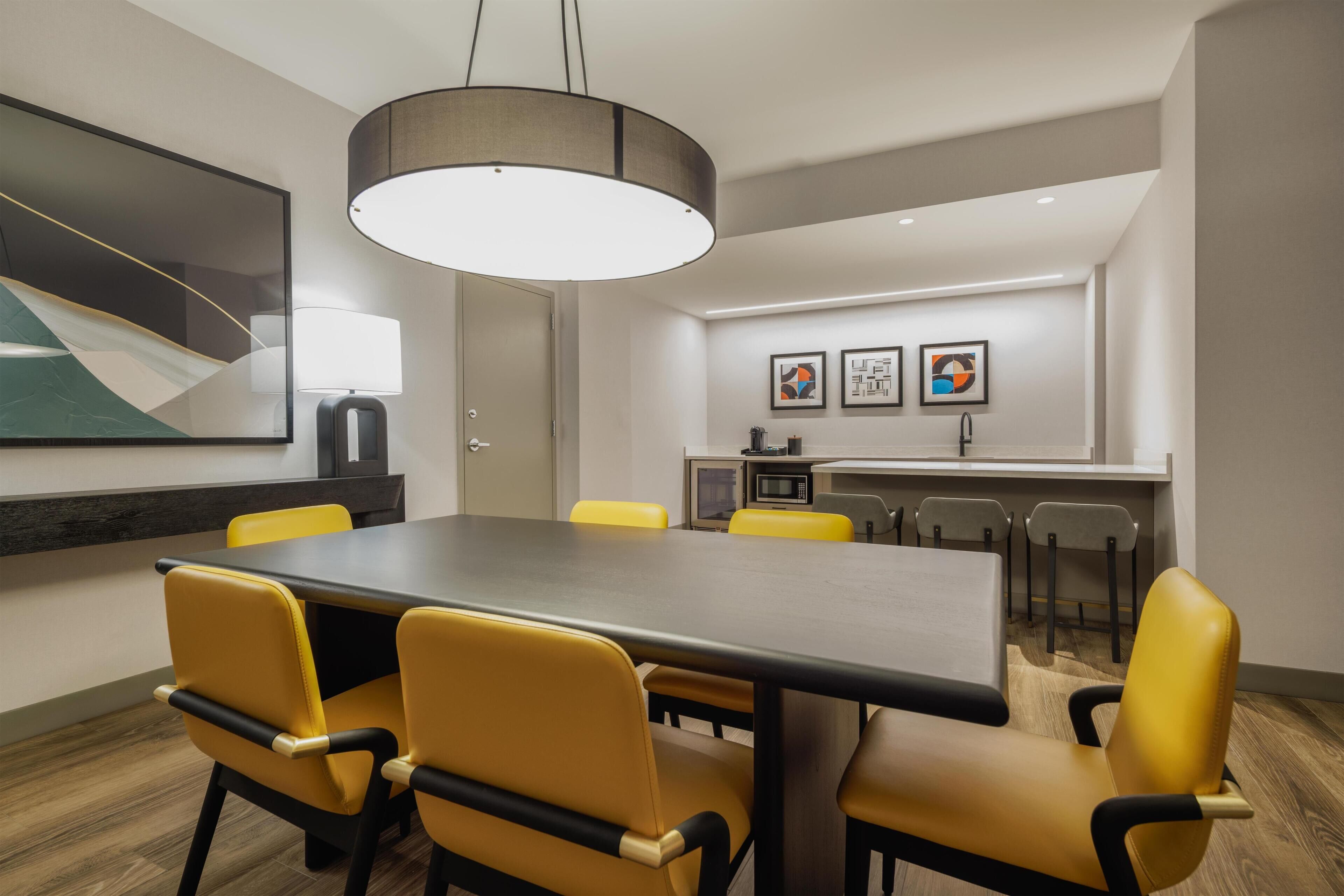 A conference room with a long table, yellow chairs, a modern pendant light, and a kitchen area in the background.