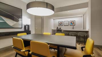 A conference room with a long table, yellow chairs, a modern pendant light, and a kitchen area in the background.