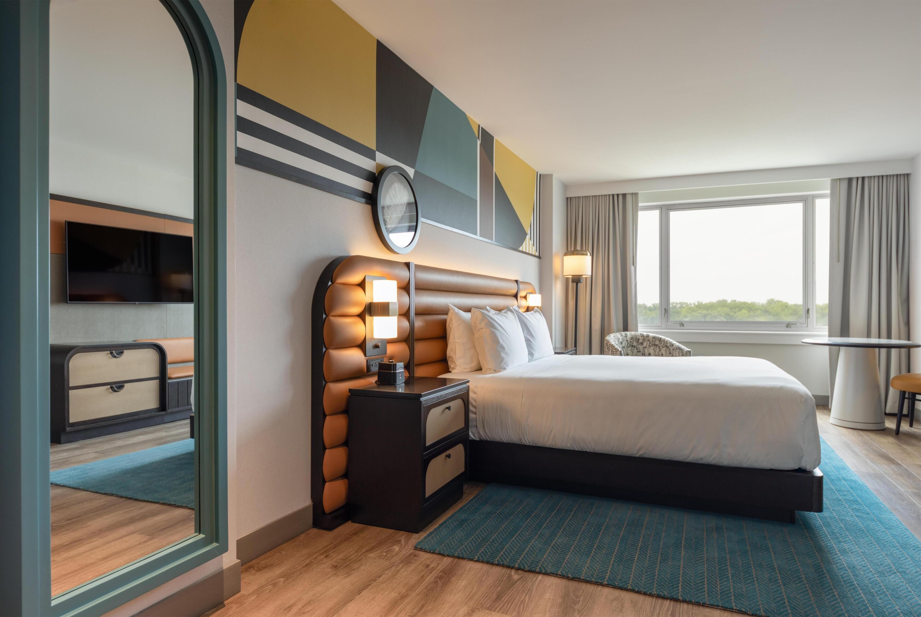 A modern hotel room with a large bed, a bedside table with lamps, a TV, and a view of the outdoors.
