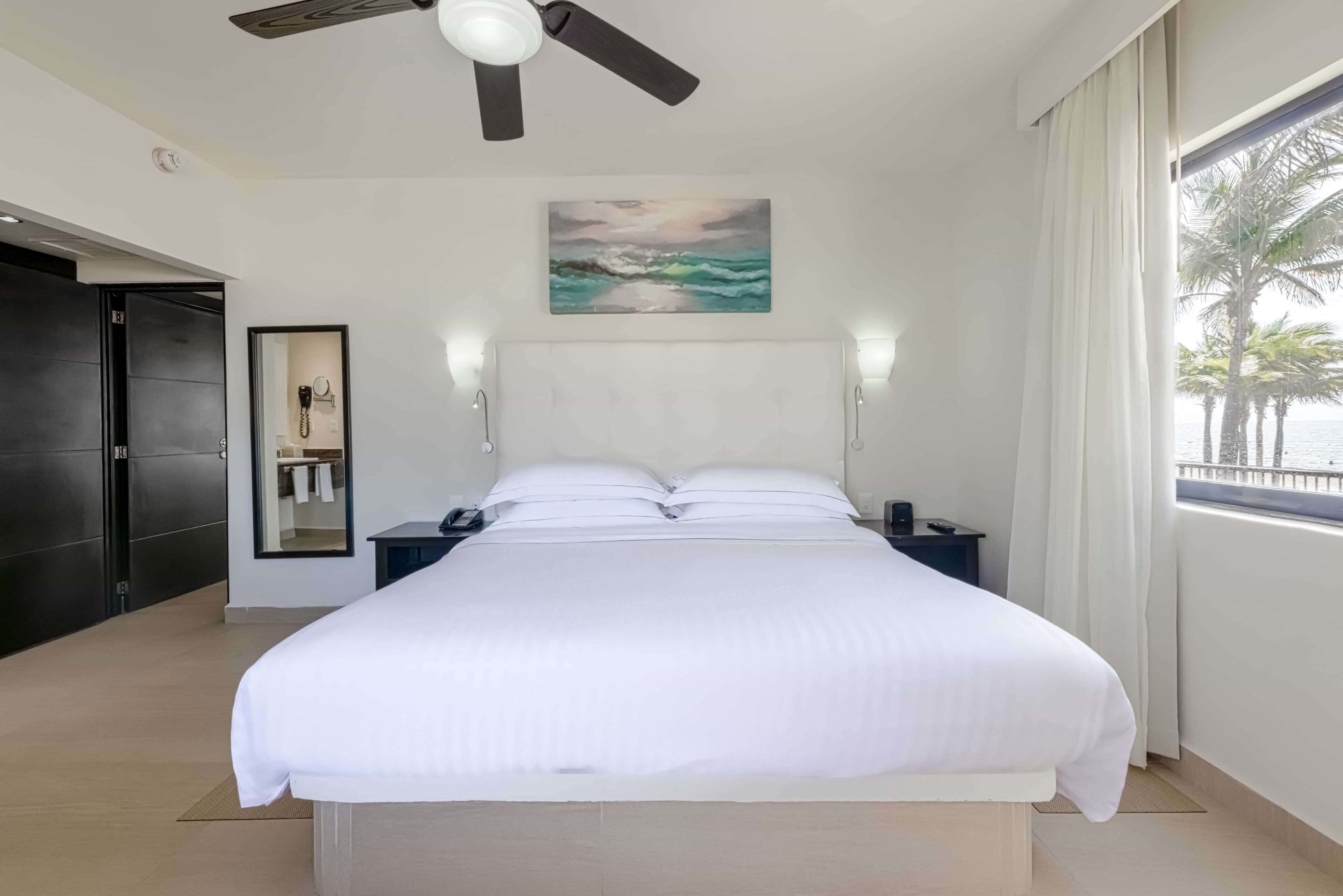 Una habitación de hotel moderna con una cama grande, un ventilador de techo, un cuadro de una playa, una mesita de noche y vistas a palmeras desde la ventana.