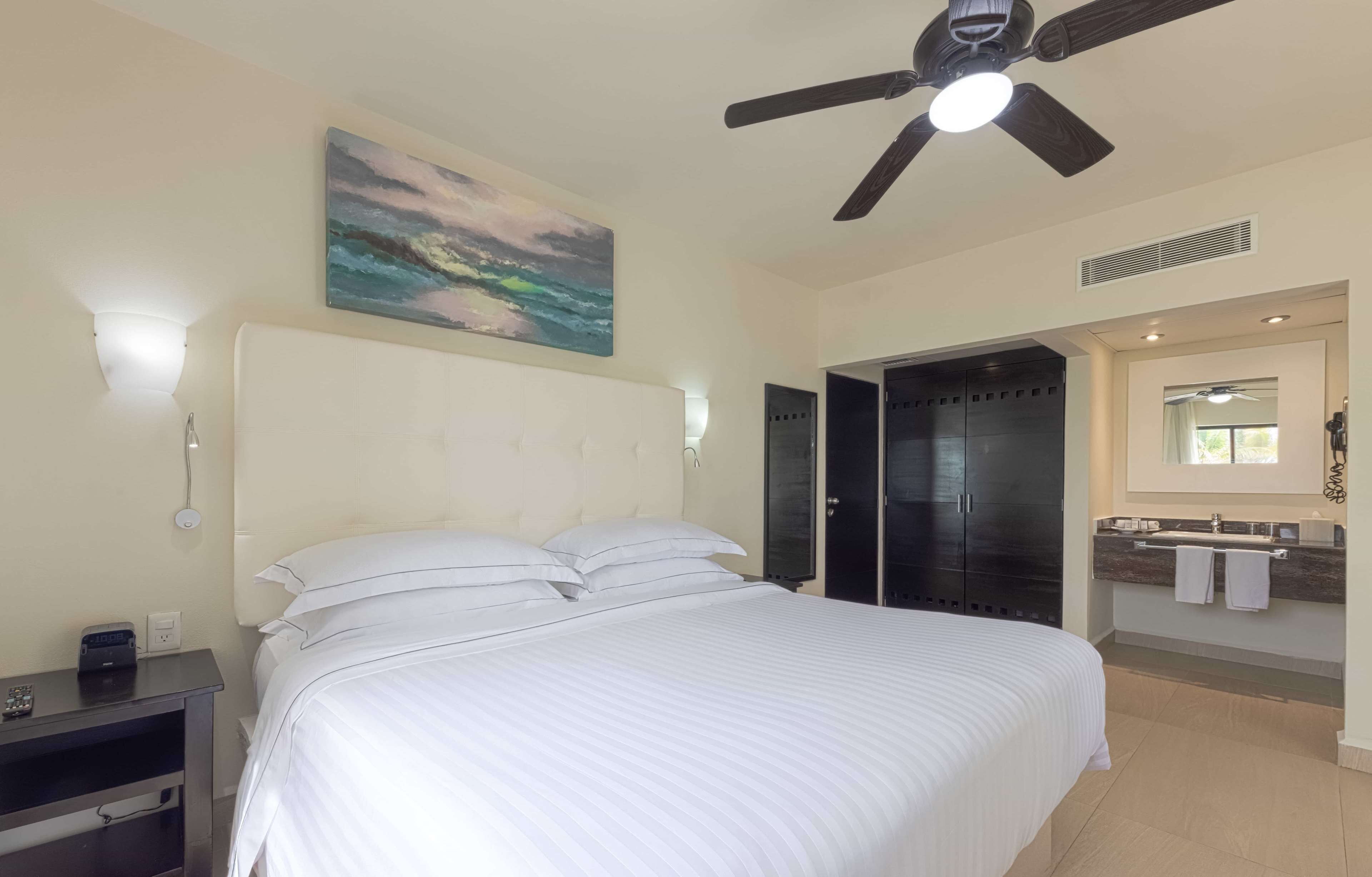 A hotel room with a bed, a ceiling fan, a painting, a desk with a lamp, and a bathroom with a mirror and sink.
