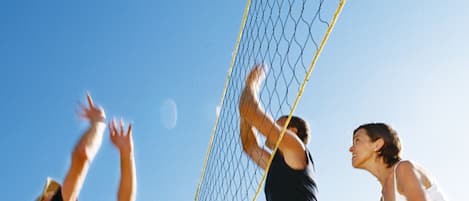 Partido de voleibol en desarrollo, con jugadores estirándose para alcanzar la pelota bajo un cielo azul despejado.