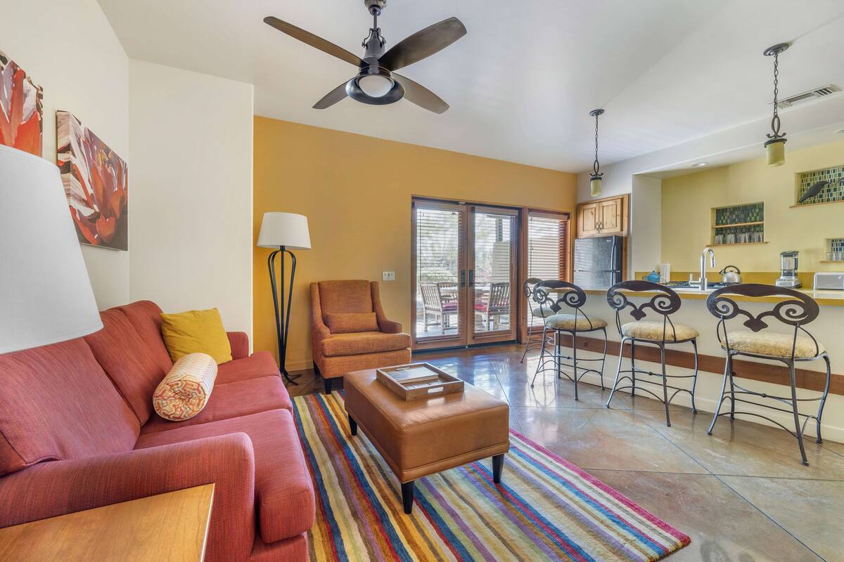 A living room with a sofa, ottoman, and a striped rug. There is a ceiling fan, a chair, and a kitchen area with bar stools.
