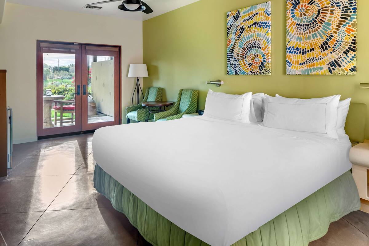 A hotel room with a large bed, a green wall, two abstract paintings, a sliding glass door leading to a patio with a chair and table, and a lamp.