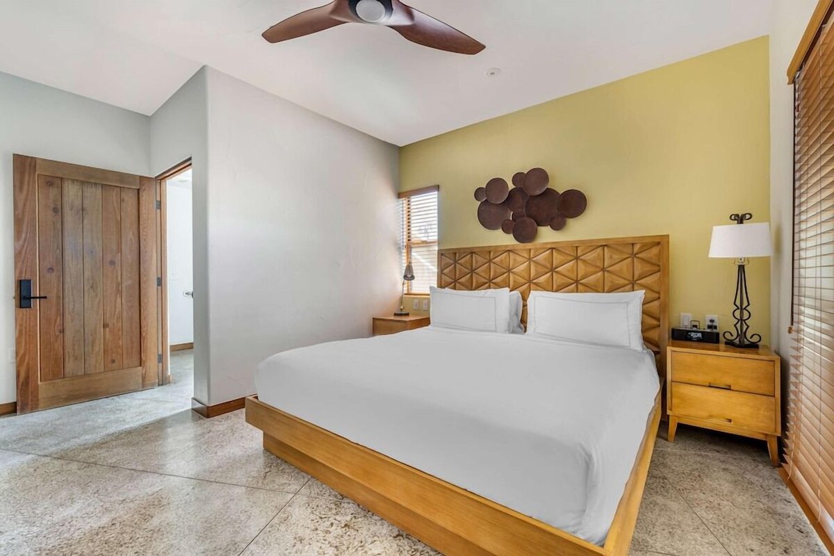 Una habitación de hotel con una cama grande, cabecera de madera, ventilador de techo, mesita de noche con lámpara y un adorno en la pared.