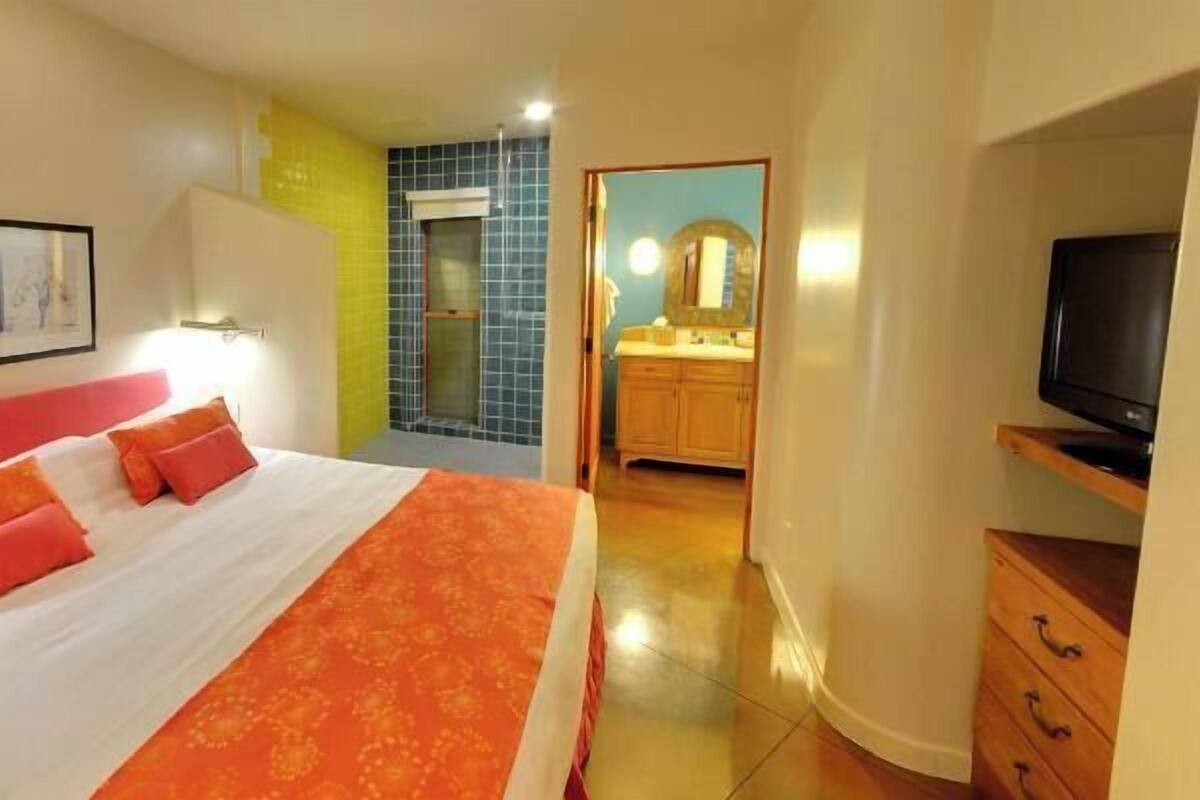 A hotel room with a bed, a bathroom with a shower, a TV, and a framed picture on the wall.