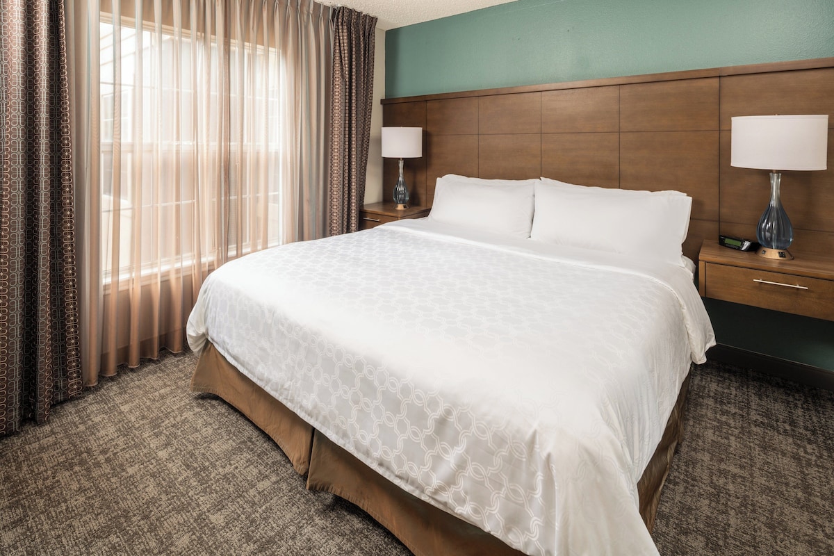 Camera d'albergo con un letto grande, due comodini con lampade, una testiera in legno e vista sull'esterno attraverso tende leggere.