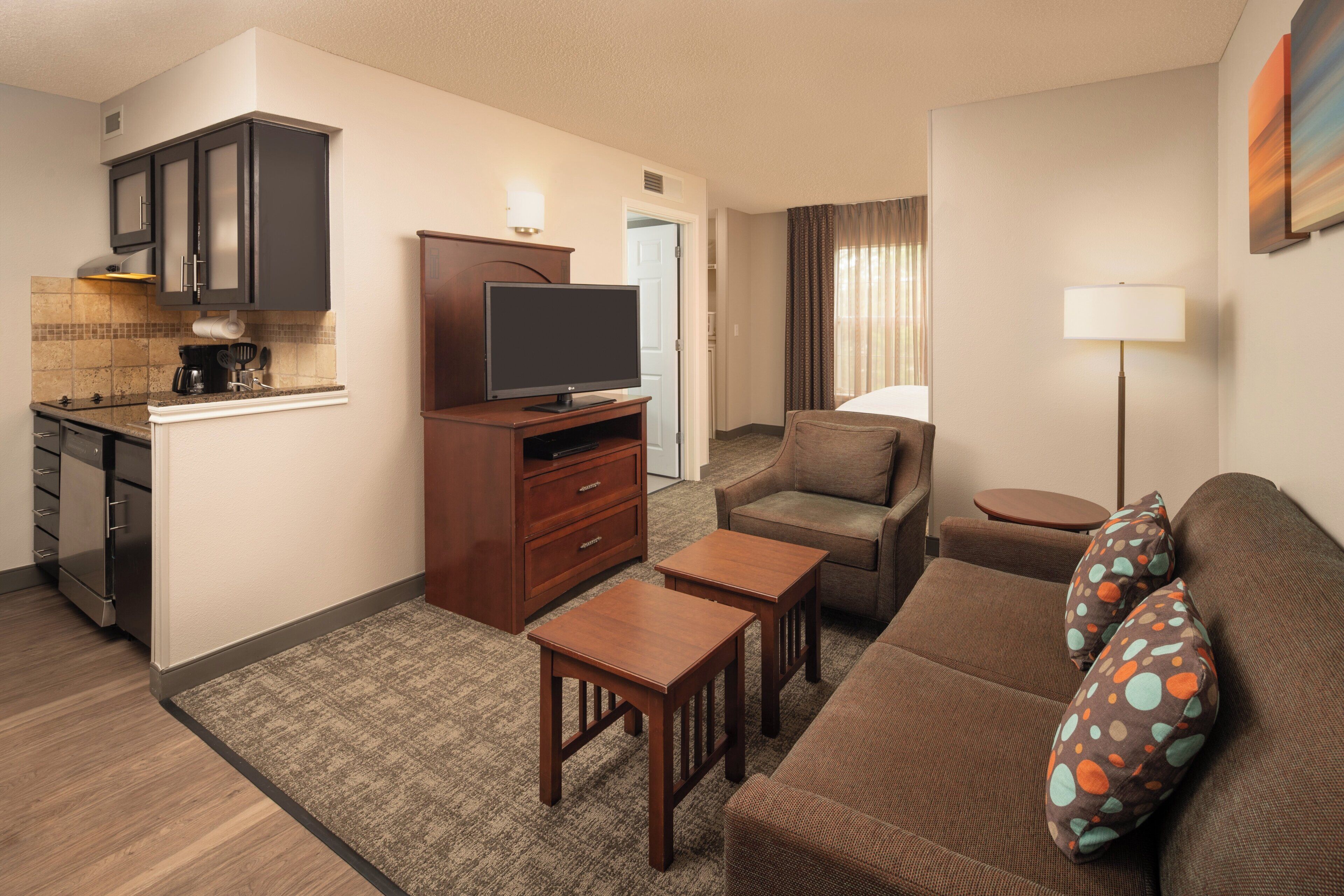 Habitación de hotel compacta con kitchenette, televisor de pantalla plana, sofá, una mesita y una lámpara.