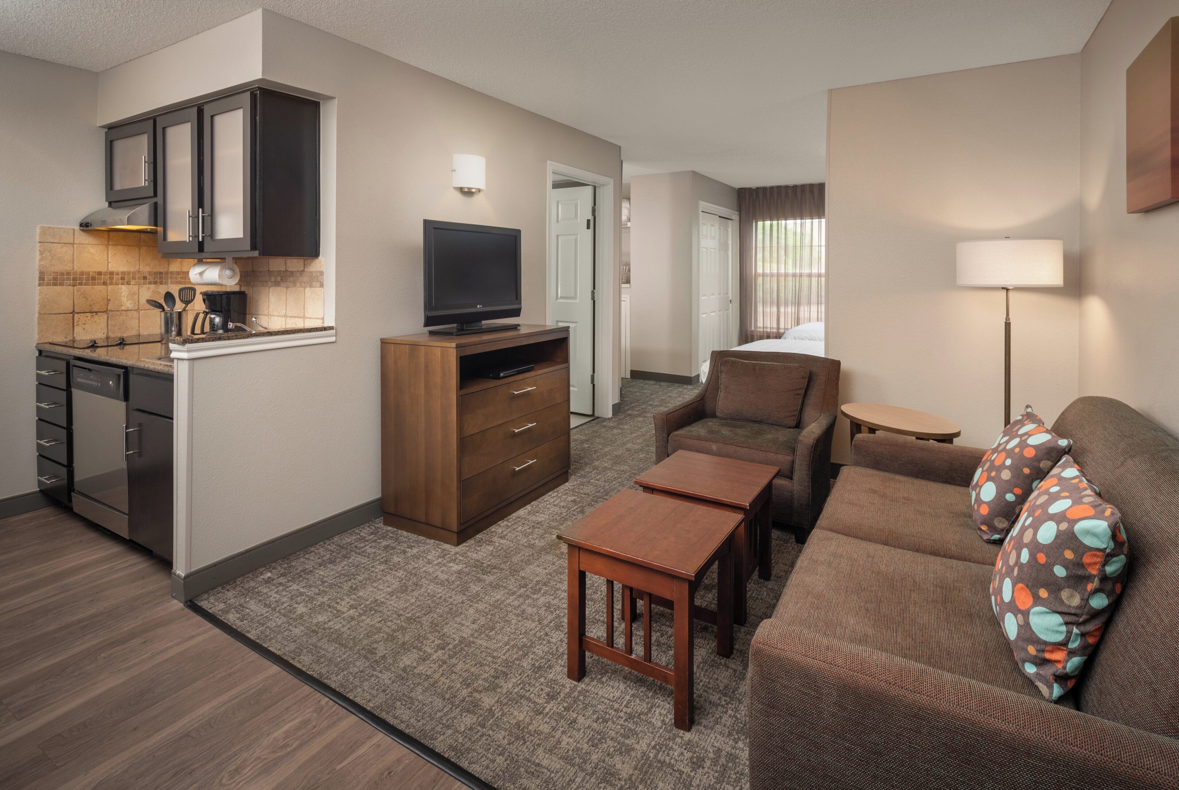 A compact hotel room with a kitchenette, a sofa, a small table, and a television.