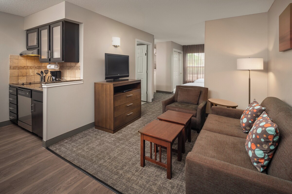 A compact hotel room with a kitchenette, a sofa, a small table, and a television.