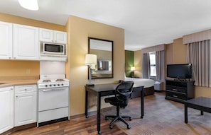 Une chambre d’hôtel compacte comprenant un coin cuisine, un lit, un bureau, une chaise, un téléviseur et une fenêtre avec des rideaux.